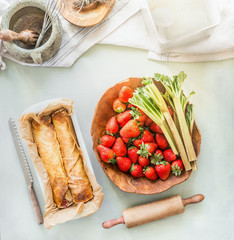 Strawberry rhubarb wrap cake on kitchen table background with bowls with ingredients. Top view. Summer baking with seasonal products . Home cuisine