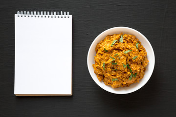 Homemade Chicken Tikka Masala in a white bowl, blank notepad on a black background, top view. Flat lay, overhead, from above.