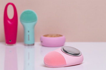 Beauty and skincare concept. Selective focus on a modern pink electric face cleansing silicone brush with massage sonic vibration on a white table and three further blurred electric beauty treatment 