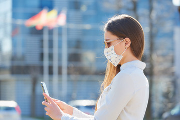 A woman using a smartphone wears a medical face mask to avoid the spread of coronavirus on a city...