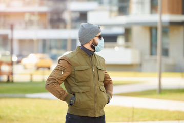 A man in a medical face mask to avoid the spread coronavirus waiting in the cozy street. A guy dropping his hands in his pockets wears a cap, sunglasses, and a face mask against COVID 19.