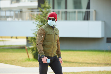 A man in a medical face mask to avoid the spread coronavirus holding his smartphone in a cozy street. A guy parading wears a red cap, sunglasses, and a face mask against COVID 19.