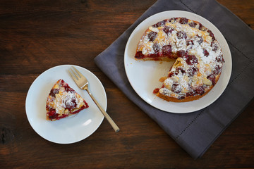 Piece and whole juicy cherry cake with icing sugar and almond slivers on white plates and dark rustic wood, high angle view from above, copy space