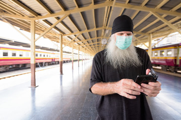 Mature bearded tourist man using phone with mask for protection from corona virus outbreak at the railway station