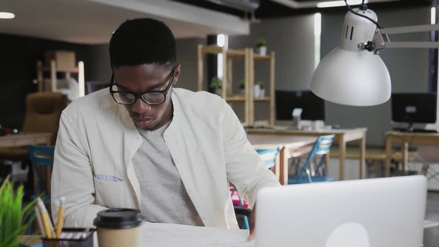 Young good looking African man in glasses sitting at the office table and working with documents