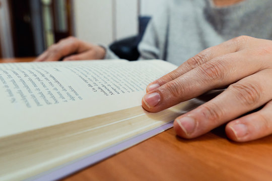 Detail of middle-aged white woman's hands picking up a book on a wooden table with coffee in the background.She is wearing a gray sweater.