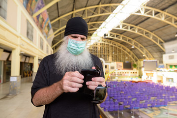 Mature bearded tourist man using phone with mask for protection from corona virus outbreak at railway station