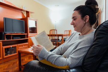 Middle-aged white woman reading a book lying on a black sofa with a yellow cushion in her spacious living room. She is wearing a gray sweater.