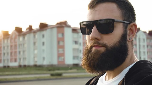 Portrait of bearded brutal image guy in black glasses. Fashionable stylish man is at sunset in city stands still in slow motion. Trendy fancy person with earring in ear. Chic, modish. Steadicam shot