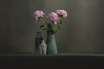Pink peonies in a vintage pottery vase and two empty small vases on a table in a grey room.