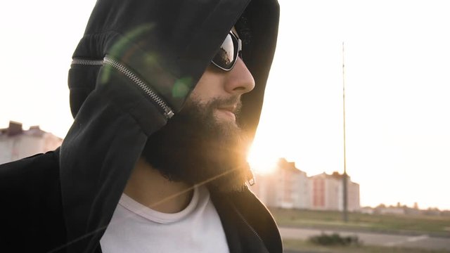 Brutal stylish man smiles at sunset in city stands still in slow motion. Bearded image modish guy in black glasses and hood. Fashionable vogue person. Concept of trendy modish people. Life style