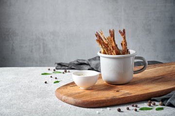 Fried smelt in grey enameled mug on brown wooden chopping board with white sauce in small sauce bowl standing on light grey textured background with dark grey cloth, side view