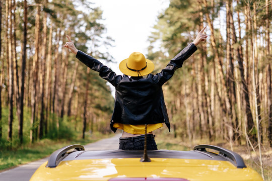 Girl Standing out of Yellow Car Sunroof Back View. Free Woman Raise Up Arms Wearing Leather Jacket and Hat. Happy Female Traveler Enjoy Forest Nature from Moon Roof. Freedom Roadtrip Concept