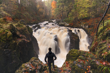 A man standing on a cliff covered with red leaves,  watches with satisfaction a beautiful waterfall of the Hermitage, Scotland in Autumn. 