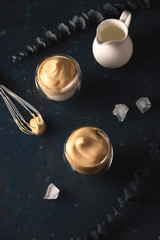 Homemade dalgona coffee made of instant coffee, sugar and milk on a dark background with ice.
