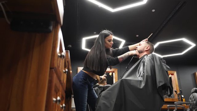 Barber woman cuts man's beard with a trimmer in barbershop. Men's hairstyling and hair cutting in salon. Grooming the hair with clipper. Hairdresser doing haircut in retro hair salon.