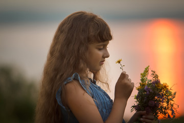 a pretty girl in a blue blouse collects a bouquet of flowers on the lake shore. sunset in the background. portrait. close up.