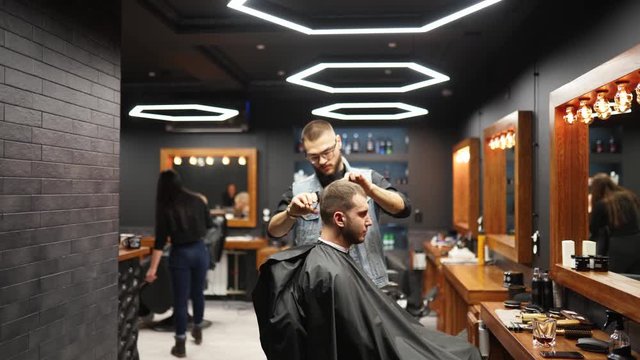 Barber guy cutting man's hair with scissors and comb. Haircutter working serving client. Stylish man sitting in vintage barber shop chair. Hairstylist man doing trendy haircut.