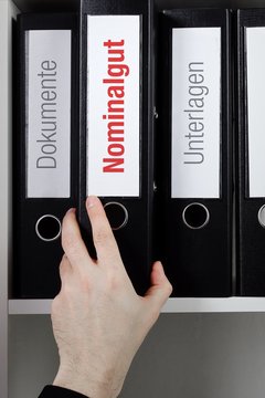Nominalgut &ndash; Finanzen/Statistik. Ordner im B&uuml;ro-Regal. Hand greift Unterlagen im Schrank. Beschriftung mit Wort