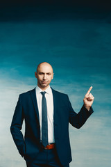 Young bald male businessman in a blue suit raised index finger up on a blue background.