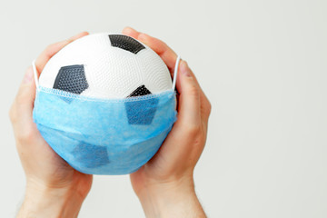 Closeup of man hands holding soccer ball in medical mask on light background during coronavirus....