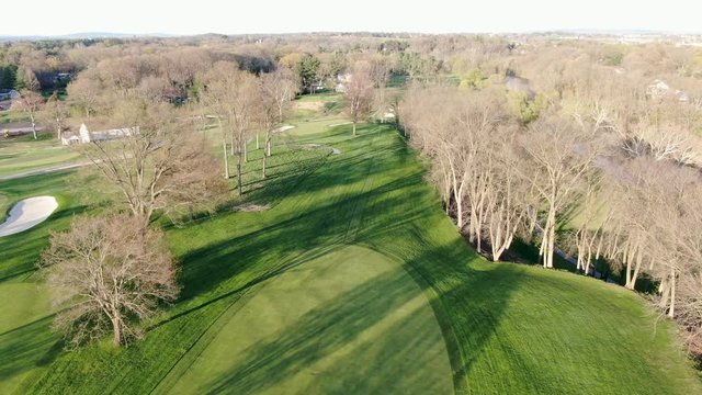 Rising aerial shot reveals golf course, green, fairway, sandtrap and water hazards, spring season, no leaves on trees