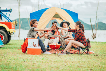 Two Asian women toasting bottles of beer to each other to celebrate a good time with a group of friends while traveling on a camping tent on holiday.