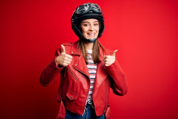 Young beautiful brunette motocyclist woman wearing motorcycle helmet and red jacket success sign doing positive gesture with hand, thumbs up smiling and happy. Cheerful expression and winner gesture.