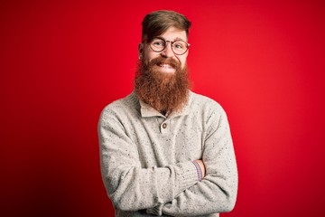 Obraz premium Handsome Irish redhead man with beard wearing casual sweater and glasses over red background happy face smiling with crossed arms looking at the camera. Positive person.