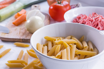 Macaroni pasta in a white bowl with other ingredients to prepare pasta bolognesa in background. Italian cuisine. Close up view.