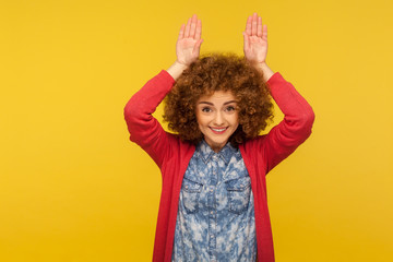 Portrait of funny positive playful woman with fluffy curly hair making bunny ears gesture, having fun and smiling with childish carefree expression. indoor studio shot isolated on yellow background