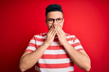 Young handsome man wearing casual striped t-shirt and glasses over isolated red background laughing...