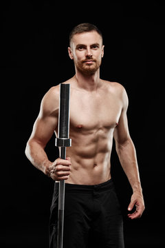 Strong athletic man - crossfit athlete fitness model showing his perfect body isolated on black background with copyspace. Holds a sports bar next to him, close-up, perfect abs, shoulders and chest