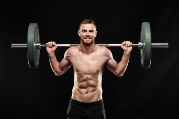 Fototapeta na wymiar Strong smiling athletic man - crossfit athlete fitness model showing his perfect body isolated on black background with copyspace. Does an exercise, holds barbell on shoulders, perfect abs and chest