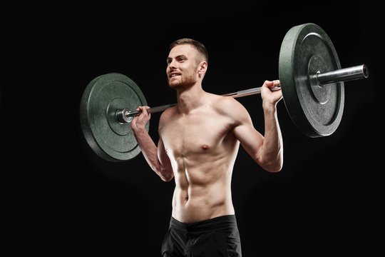 Strong smiling athletic man - athlete fitness model showing his perfect body isolated on black background with copyspace. Does an exercise, holds barbell on shoulders, perfect abs and chest