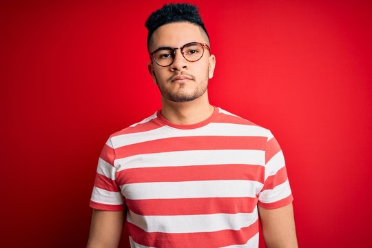 Young handsome man wearing casual striped t-shirt and glasses over isolated red background Relaxed with serious expression on face. Simple and natural looking at the camera.
