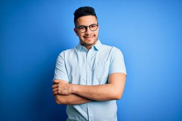 Fototapeta premium Young handsome man wearing casual summer shirt and glasses over isolated blue background happy face smiling with crossed arms looking at the camera. Positive person.