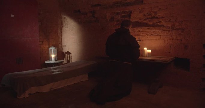 Monk gets on knees, starts praying in front of altar in dark room. Silhouette, candle light