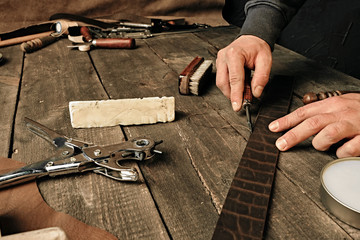 Working process of the leather belt in the leather workshop. Man holding tool. Tanner in old tannery. Wooden table background. Close up man arm. Maintenance concept. Goods production.