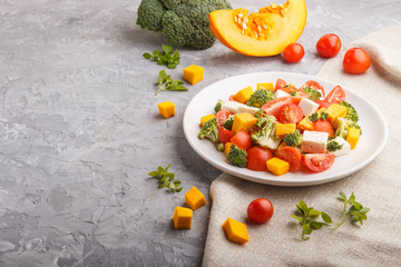 Vegetarian salad with broccoli, tomatoes, feta cheese, and pumpkin on white ceramic plate on a gray concrete background, side view, copy space.
