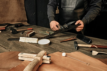 Working process of the leather belt in the leather workshop. Man holding tool. Tanner in old tannery. Wooden table background. Close up man arm. Maintenance concept. Goods production.