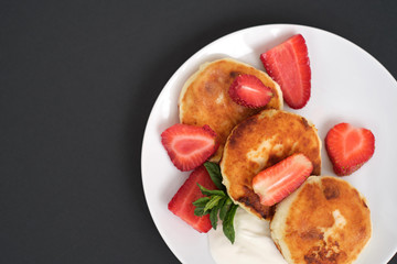Russian and Ukrainian cuisine. Breakfast. Cottage cheese pancakes. Cheesecakes with fresh strawberries, mint and sour cream sauce in a white plate on a light grey background. Copy space close-up 