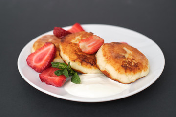 Russian and Ukrainian cuisine. Breakfast. Cottage cheese pancakes. Cheesecakes with fresh strawberries, mint and sour cream sauce in a white plate on a light grey background. Copy space mocap close-up