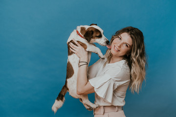 Adorable blonde in casual dress, broad smiling, holds her puppy Jack Russel, who is licks her cheek, over blue background. Pets and hosts. Happiness concept.