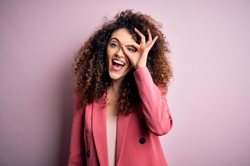 Young beautiful businesswoman with curly hair and piercing wearing elegant jacket doing ok gesture with hand smiling, eye looking through fingers with happy face.