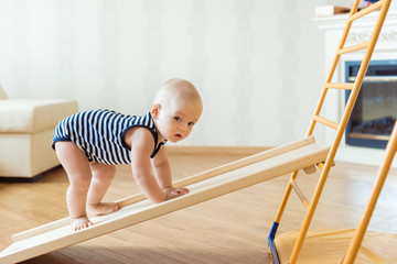 Cute baby performs gymnastic exercises on a wooden home sports complex stairs and rings. Children's...