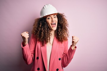 Young beautiful architect woman with curly hair wearing safety helmet over pink background celebrating surprised and amazed for success with arms raised and open eyes. Winner concept.