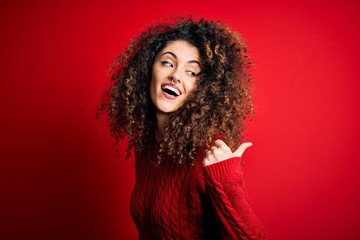 Young beautiful woman with curly hair and piercing wearing casual red sweater smiling with happy face looking and pointing to the side with thumb up.