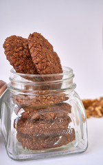Open lid glass cookie jar with oatmeal cookies inside with walnuts in the background on white surface table - delicious gluten-free oatmeal cookies with walnuts and cocoa