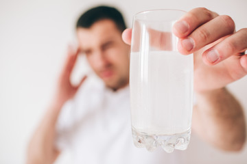 Young sick man isolated over background. Guy hold glass of water with effervescent pills and medicine in it. Suffer from headache and pain. Blurred background.